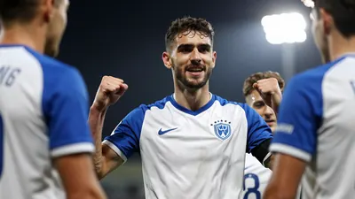A focused Gent player in a blue and white kit, celebrating a goal with teammates