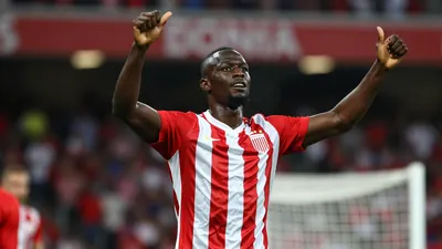 Muscular African striker in a red and white striped jersey, mid-celebration after scoring a goal, arms raised, face lit...