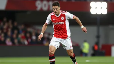 A photorealistic image of a Gent player in a red and white jersey, with a white kit and a black and white striped sock...
