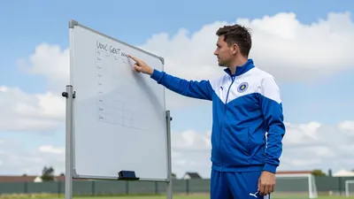 A 30-year-old Gent assistant coach, wearing a blue and white tracksuit, stands on the training pitch, looking at a...