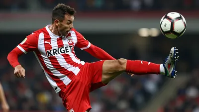 A dynamic action shot of a KRC Gent player in a red and white striped jersey, mid-kick, with the ball in the air, taken...