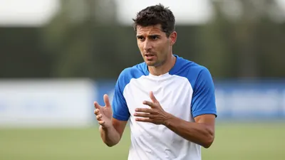 Mid-30s male coach, athletic build, intense eyes, short dark hair, slight stubble, waist-up in a plain blue and white...