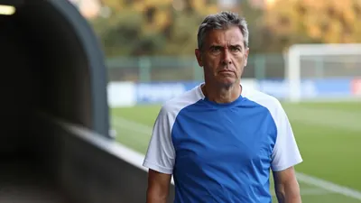 A 50-year-old, 5'9" Gent coach with a stern expression, wearing a plain blue and white polyester training top, stands on...